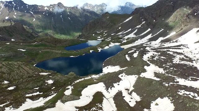 Aerial view of alpine mountain lake surrounded by melting spring snow and rugged peaks. High elevation tundra green meadow landscape create breathtaking remote wilderness scenery.