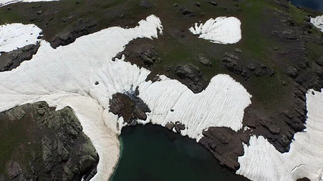 Aerial view of alpine mountain lake surrounded by melting spring snow and rugged peaks. High elevation tundra green meadow landscape create breathtaking remote wilderness scenery.