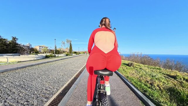 Woman bicycle coast young woman in a red backless jumpsuit cycles along a scenic paved path by the bright blue ocean