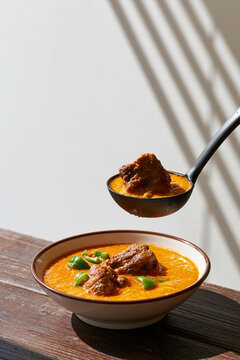 Close-up of curry with meat and green peppers in a bowl with ladle under dramatic window light.