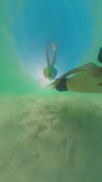 Vertical video. Underwater Scene Diver Swimming Along Sandy Bottom Wearing Mask With Woman In Inflatable Ring Above, Sunlight Filtering Through Water, Wide Angle Perspective, Slow Motion Exploration