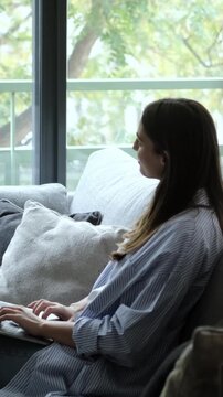 Side view of a confident young businesswoman typing on her laptop keyboard, comfortably sitting on a cozy sofa next to a large window in her modern home office while working remotely