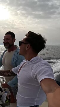 Joyful young man wearing sunglasses filming himself on a moving motorboat with his friend steering, enjoying a summer sea adventure and sharing the experience on social media