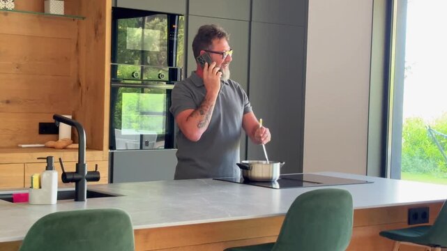 Busy multitasking man with a beard and glasses talking on the phone while stirring food in a pot on an induction stove, staying connected while preparing a meal in his modern home