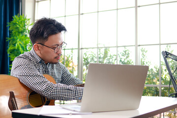 Fototapeta premium Asian male musician writing lyrics and playing acoustic guitar in home studio with microphone and laptop. Concept of songwriting, music production, and podcasting.