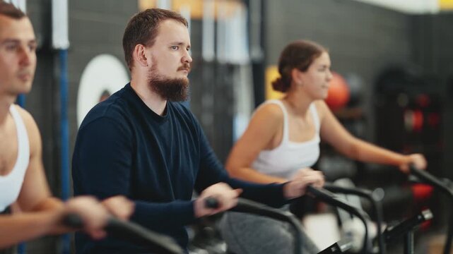 Focused motivated young adult man leading healthy active lifestyle doing cardio workout on air bike during group cross training in gym