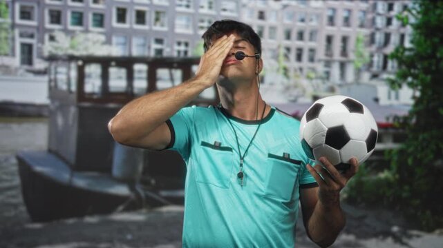 Man referee holding soccer ball, hand to forehead in front of building wearing whistle and headset, turquoise uniform, eyes closed; frustration defeat.