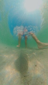 Vertical video. Underwater Low Angle View Couple Walking On Sandy Bottom With Strong Bubble Splash Above, Legs Moving Through Clear Water, Wide Angle Distortion, Tropical Seascape