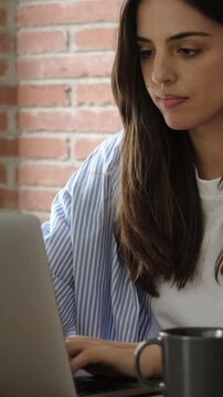 Confident young businesswoman working on her laptop in a modern office with a brick wall, focused on her task while typing on the computer keyboard and drinking a cup of coffee