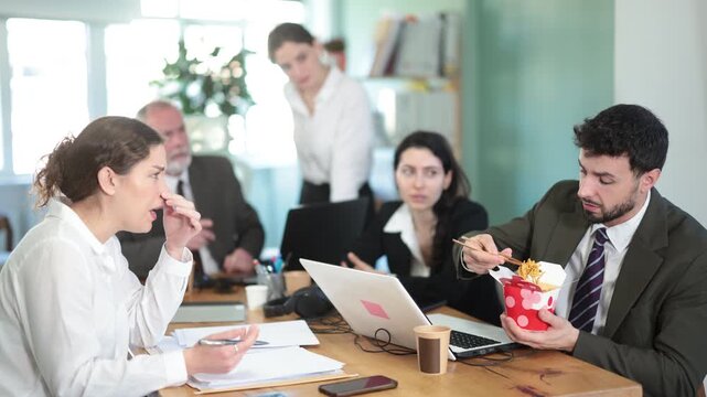 Positive young man office worker eating food at workplace, while other co-workers unhappy and continue to work in bright office of large business company