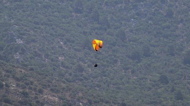 Two yellow orange Spanish paragliders glide down beside a scrub forest mountain slope. Pair of colorful pilots descend near rugged hillside vegetation in Spain adventure scenery.