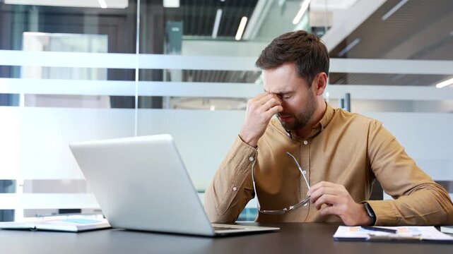 Overworked businessman suffering eye strain and headache while working on laptop at desk in office, taking off glasses and rubbing nose bridge. Fatigue, stress, digital overload during long work hours
