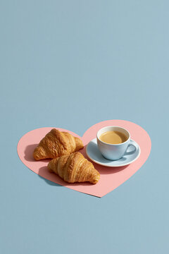 Two croissants and coffee on a pink heart-shaped paper on a light blue surface with soft shadows.