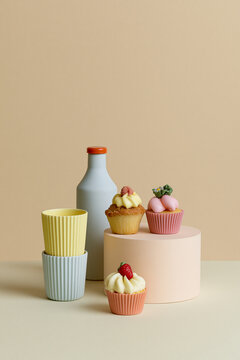 Minimalist studio arrangement of cupcakes, cups and a bottle in soft pastel tones on a beige background.