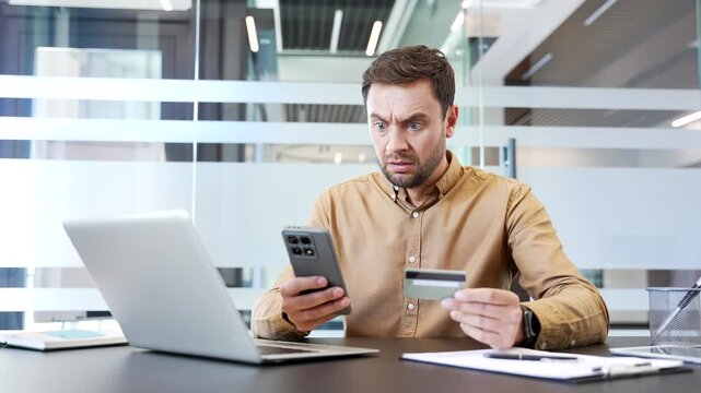 Shocked businessman checking bank details on smartphone while sitting at desk in office, reacting to suspicious charges with distress. Online fraud, financial loss and security issue in mobile banking