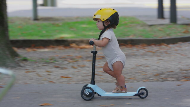 Toddler wearing yellow helmet riding blue scooter in park pathway, captured in side profile, focused and balanced while gliding forward during playful outdoor moment