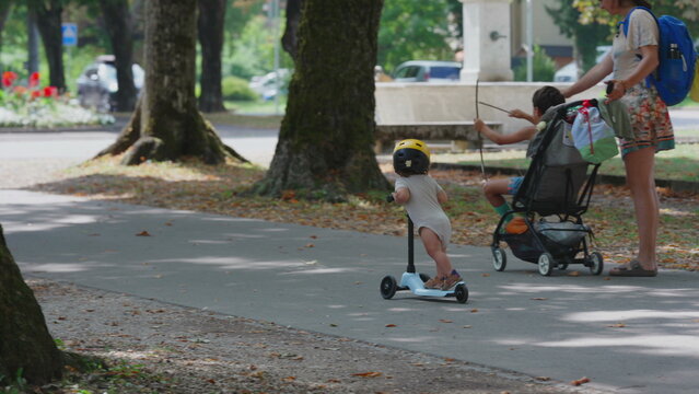 Toddler riding scooter on sunny park pathway wearing yellow helmet, distant view with parent and stroller ahead, playful and curious outdoor childhood activity