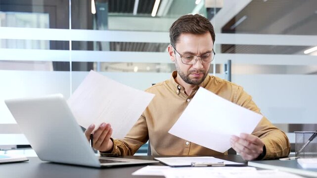 Confused businessman reviewing financial documents at desk in modern office, struggling to understand poor results. Frustration, analysis difficulty and concern during stressful business workflow.