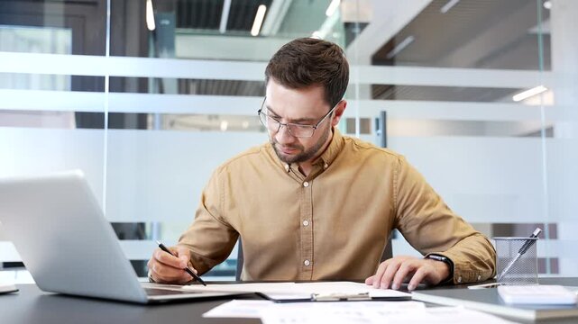 Confused businessman reviewing documents at desk in office, unable to understand paperwork, looking at camera with puzzled expression. Work difficulty, misunderstanding, problem situation in workflow.