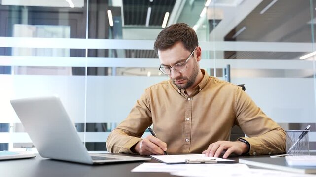 Businessman sitting at desk in office examining contract, signing document, finalizing agreement. Paperwork completion, legal confirmation, business deal execution in professional corporate workflow