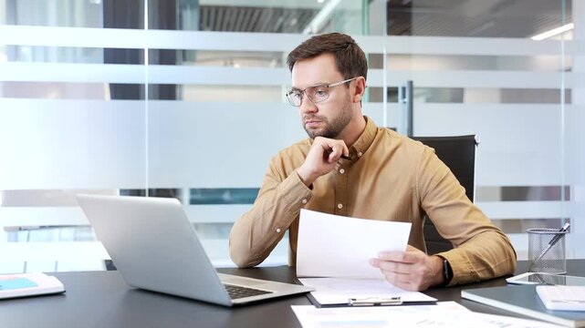 Thoughtful businessman working on laptop, holding document at desk in office, comparing data and checking details. Focused review, verification process and careful decision making in business workflow