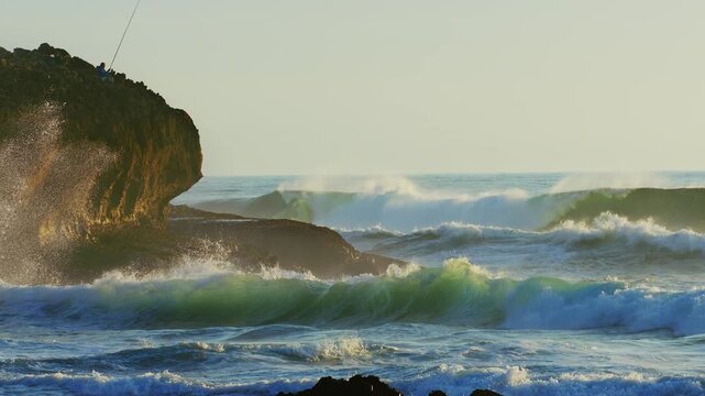 Cinematic 4K slow motion footage of powerful ocean waves crashing against a rugged rocky cliff where a lone fisherman stands. The scene captures the raw beauty of the coastal landscape bathed in the s