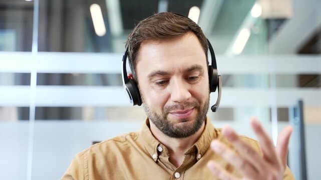 Close up webcam view of smiling businessman wearing headset speaking on video call sitting in office, looking at camera. Coaching session, knowledge sharing and online training in corporate workflow.