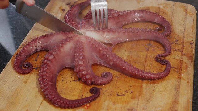 Chef is cutting a freshly boiled red octopus with a sharp knife on a wooden board. Shows the process of preparing exotic seafood for a gourmet meal.