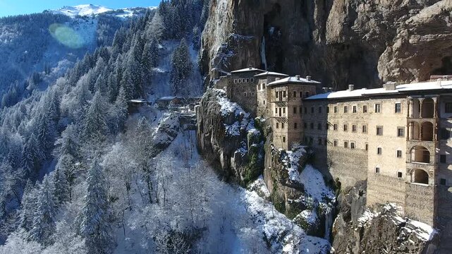 Aerial drone view captures snow covered Sumela Monastery clinging to steep rocky mountain cliff. Forested valley shows valley slope, winter season, and ancient christian plus christian site.