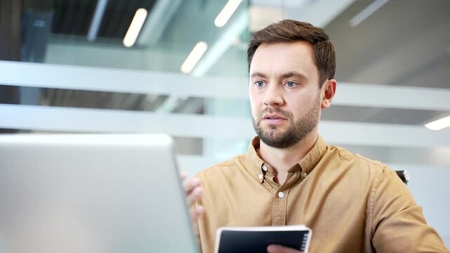 Close up of businessman on video call using laptop while sitting at desk in modern office, speaking and taking notes. Remote training, online learning and professional communication in daily workflow.