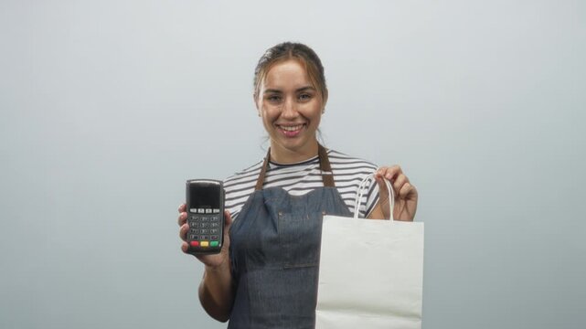 Young blonde woman wearing denim apron and striped shirt holding a dataphone and white paper bag while sticking tongue out in studio; playful promotion.