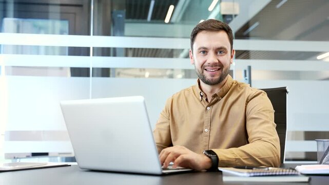 Portrait of smiling businessman at workplace desk using laptop in modern office, looking at camera with confident expression. Positive mood, daily workflow, professional in corporate environment.
