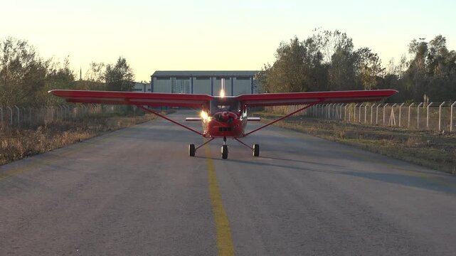 Red single engine airplane taxiing from hangar toward airstrip on quiet airport runway. Light trainer plane rolls slowly before takeoff with propeller spinning in calm morning light.