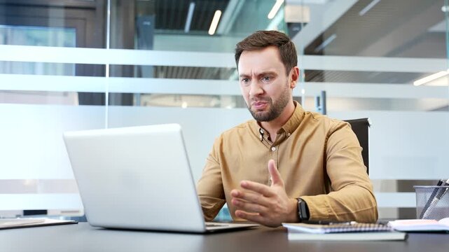 Angry businessman arguing on video call using laptop while sitting at desk in modern office, expressing frustration during online meeting. Conflict situation, disagreement and intense communication