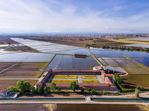 Aerial view of Tenuta Colombara historic farm estate surrounded by flooded rice fields under a clear blue sky in Livorno Ferraris, Piemonte, Italy.