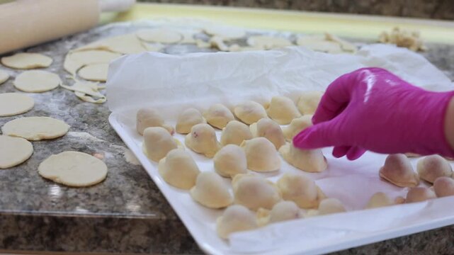 hand in pink glove arranging raw homemade dumplings on white parchment tray with dough circles rolling pin and flour on counter