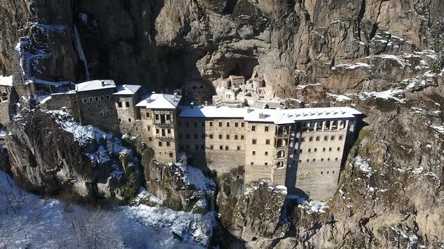 Aerial drone view captures snow covered Sumela Monastery clinging to steep rocky mountain cliff. Forested valley shows valley slope, winter season, and ancient christian plus christian site.