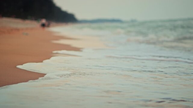 Beautiful sea waves with white foam washing onto golden sandy beach at sunset. Low-angle shoreline view captures a peaceful nature atmosphere, ideal for summer travel, vacation, and wellness concepts.