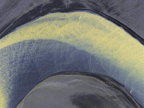 Aerial view of the abstract patterns of a glacial river with yellow sediment mixing with blue water over dark volcanic sand in Hallgeirseyjarvegur, Rangarthing eystra, Iceland.