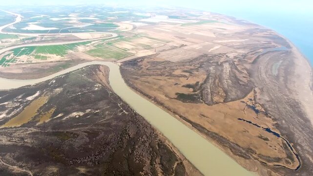 Panoramic aerial view of longshore drift sands shaping river mouth on coastal alluvial plain. Sediment accumulation creates a sand spit barrier where channel meets sea along flat delta terrain.
