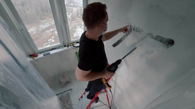 Worker drilling horizontal hole in wall near window on ladder