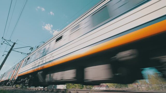 Suburban Electric Train Passing at High Speed: Low Angle Bottom-Up Close-Up with Motion Blur | White Orange Railway Cars, Track and Overhead Wires on Clear Blue Sky