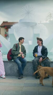 Two happy high school friends are seen talking and laughing while sitting by a fountain in a city park. They are enjoying their casual conversation and drinks outdoors during their free time.