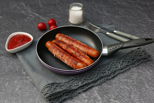 Three fried Bernese sausages in a pan with ketchup, cherry tomatoes and cutlery on a dark stone background.