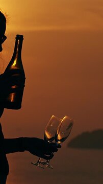 Vertical video. Side Silhouette of Woman Holding Champagne Bottle and Two Glasses against Bright Sunset over Ocean Warm Golden Light Romantic Evening Leisure and Travel Lifestyle Scene
