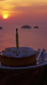 Vertical video. Close View of Birthday Cake with Candle on Plate against Blurred Ocean Sunset Background Warm Evening Light Celebration and Travel Lifestyle Scene
