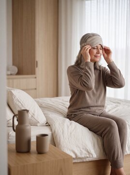 Mature woman sitting on bed and putting on a silk sleep mask for a restful night's sleep in a minimalist bedroom