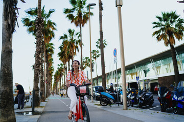 Carefree woman with braided hair riding bike under palm trees, laughing with eyes closed, embodying pure joy and light-hearted freedom on bright summer day in city. © BullRun
