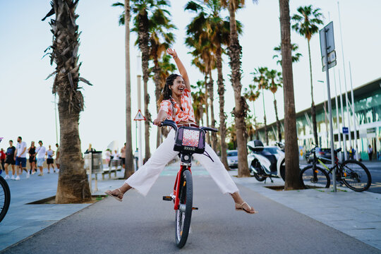 Laughing young Black woman cycling with arm raised in joy, embracing digital age mobility, feminine empowerment and vibrant lifestyle through modern shared transport.