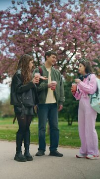Happy high school friends meet in a park and share drinks on a sunny spring day. Group of cheerful teens reconnect outdoors under beautiful blossoming cherry trees.